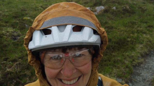 Amanda Wagstaffe taking a selfie outdoors in a mountainous, green landscape. She is smiling and wearing a white bike helmet, safety glasses, and a rain jacket with the hood up, as the weather appears misty and damp.