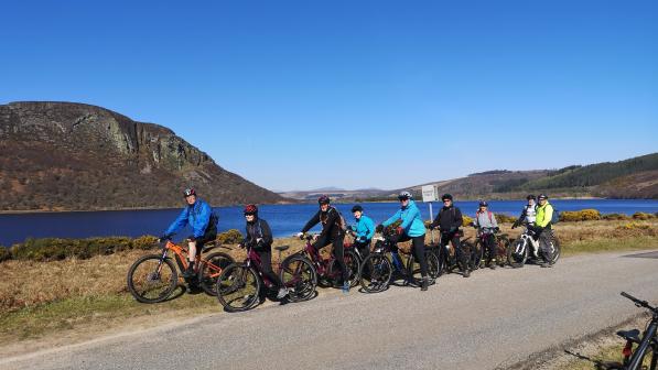 A group of people all on e-bikes are standing astride their bikes next to a quiet country single-track road with a loch and mountains in the background