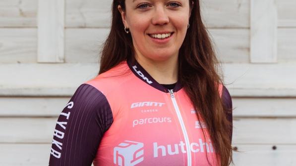 Amy Marks is standing in front of a white-painted wooden fence. She is wearing a purple and pink short-sleeved cycling jersey. She has long brown hair and cycling sunglasses on her head.