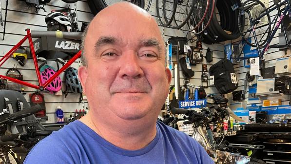 A man in a blue T-shirt is sitting in front of a wall with lots of cycling-related paraphernalia attached to it, wheels, tyres, helmets, a frame and so on