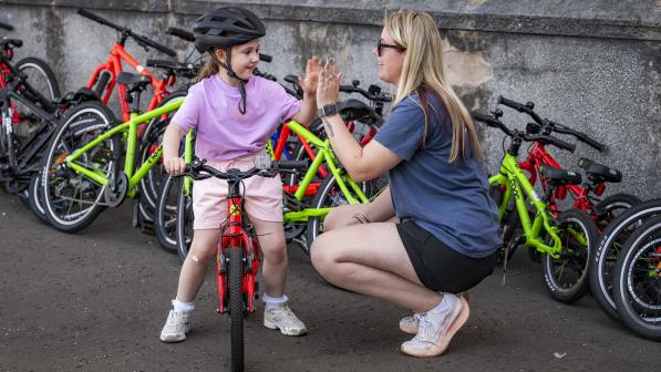 A woman with long blonde hair and a blue T-shirt is kneeling down to high-five a young child in pink T-shirt and shorts sitting on a children's bike