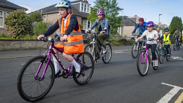 A group of school children accompanied by some adults are riding along a urban road. They're wearing a mix of school uniform and normal clothes and some are in hi-vis jackets