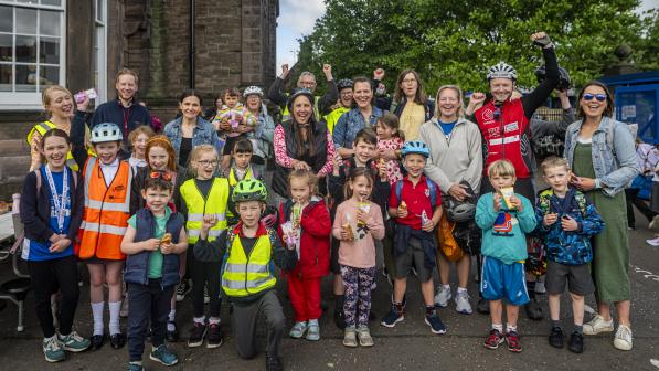 A group of parents and children stand in a school playground, cheering towards the camera after arriving at school as a Bike Bus group