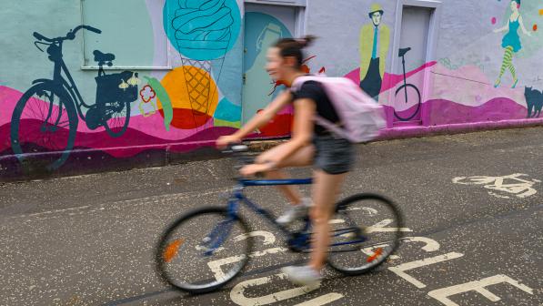 A woman is cycling on a blue bike. She's wearing shorts and T-shirt and has a pink rucksack. She's deliberately blurred so the background is in focus. It shows a colourful mural with bikes and ice cream and people
