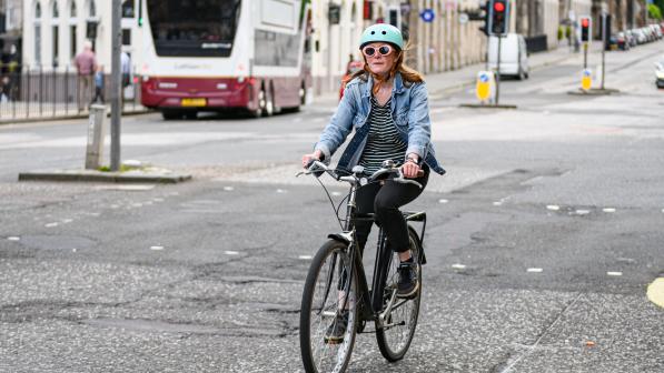 A woman is riding a black hybrid bike with a rear rack on it on an urban street. She is wearing black trousers, a stripy top and blue denim jacket. She has long red hair, a turquoise cycling helmet and sunglasses.
