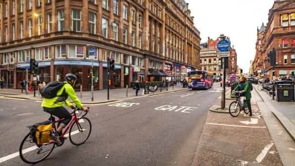 A person is cycling along an urban road towards a bus gate. There's another person on a bike waiting to turn right in front of them
