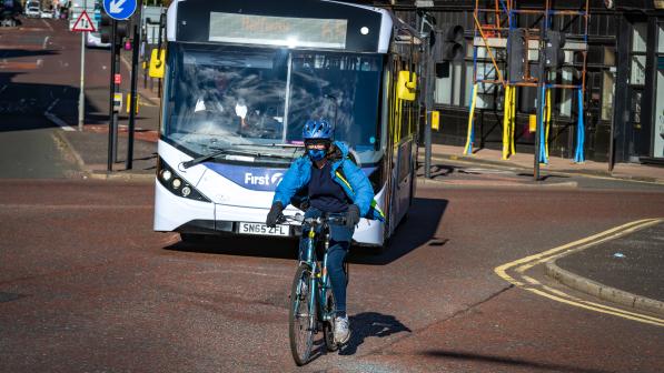 A person on a blue hybrid bike is riding along an urban road with a bus behind them. They're wearing blue jeans and top and a blue jacket, as well as a blue cycling helmet and face covering. They clearly like blue