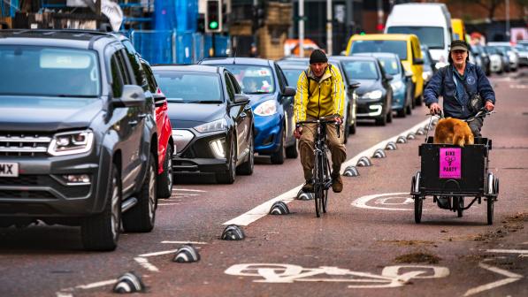 A man in trousers and a yellow jacket riding a road bike and a man in blue jacket riding a cargo bike with a dog in it are cycling along a separated cycle lane past a line of cars