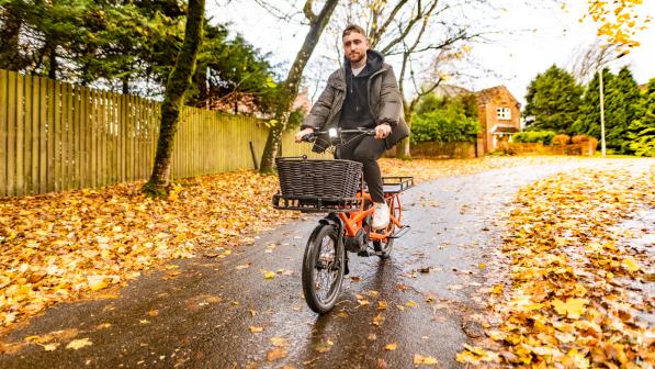 A man is cycling on a wet road that's got wet leaves to both sides. He's riding an orange cargo bike and is wearing winter clothing