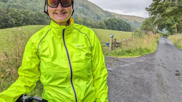 A smiling cyclist wearing a bright yellow waterproof jacket and a red helmet is standing next to her bike on a quiet country lane. The background features lush green hills, trees, and a tranquil rural landscape on a cloudy day.