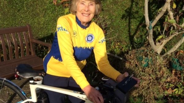 A smiling cyclist wearing a yellow and blue cycling jersey stands next to her road bike in a garden setting. The background features greenery, with a bench and plants visible, creating a warm and welcoming atmosphere. She appears ready for a ride, exuding enthusiasm and passion for cycling.