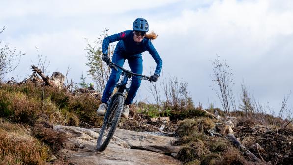 Anna Riddell is riding a mountain bike down a technical off-road trail. She is wearing blue mountain biking kit and helmet and cycling sunglasses. She is smiling.