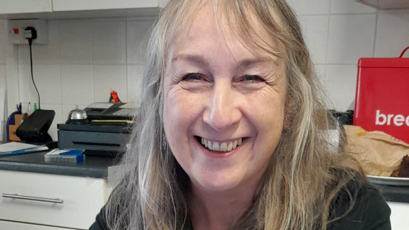 Annette Cormack is sitting at a table in a kitchen. She has a laptop in front of her. She is wearing a Community Recycle Cycles T-shirt. She has long hair and is smiling at the camera