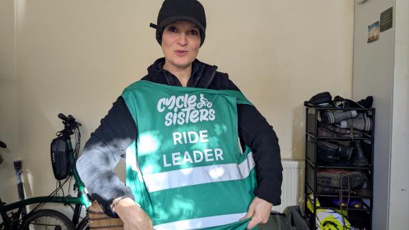 A smiling woman wearing a black cap and a green high-visibility vest with "Cycle Sisters Ride Leader" printed on it stands indoors. The background features bicycles and equipment, reflecting her involvement in cycling. Her confident and approachable demeanor highlights her leadership role in empowering others through cycling.