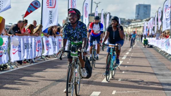 A joyful cyclist wearing a floral shirt, helmet, and glasses rides confidently along a road during an organised event. Behind them, other cyclists participate in the ride, with banners displaying "London to Brighton Skyline." The vibrant atmosphere and supportive community highlight their enthusiasm and commitment to cycling.