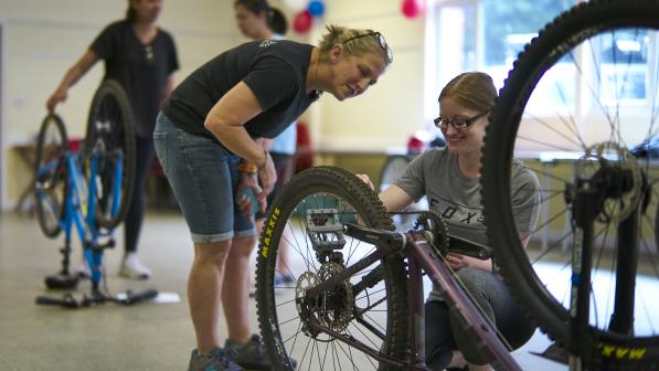 A woman crouches beside another woman who is learning to fix a bike wheel at an indoor Big Bike Revival event, with others working on bikes in the background.
