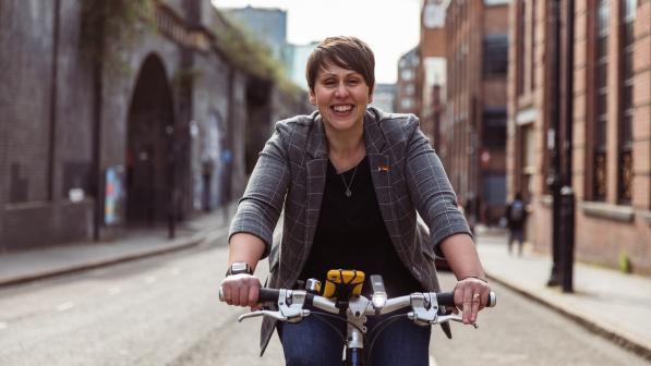 Beccy Marston is riding along an urban street. You can just see the flat handlebar of her bike. She is wearing a grey jacket with white check pattern, black top and blue jeans. There is a rainbow badge on the lapel of the jacket. Her smartphone is attached to her handlebar. She is smiling at the camera.