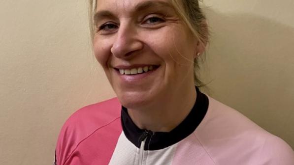 A smiling woman wearing a pink and white Kent Velo Girls cycling jersey poses against a plain background. Her friendly and approachable demeanor reflects her passion and dedication to fostering a supportive and inclusive cycling community for women.