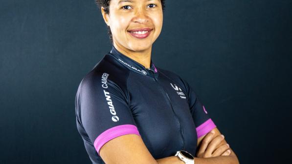 A portrait of Bianca Fernandez. She is standing with arms folded against a blue-grey background. She is wearing a dark blue cycling jersey with a multicoloured pattern around the bottom and purple cuffs.