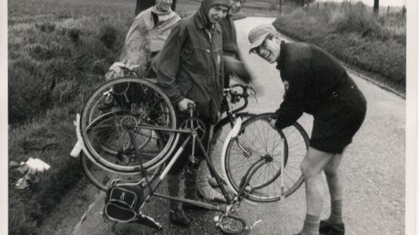 A scan of an old black and white photo showing a group of men with bikes. One bike is turned over and propped up on its saddle as one of the men takes off the front wheel