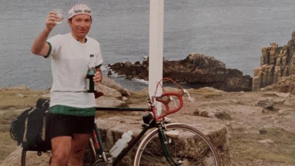 A man in white T-shirt and black shorts is standing in front of a loaded touring bike and holding a bottle and a glass which he has raised to the camera. There are cliffs and the sea in the background. The bike is leaning against a white sign reading 'Land's End 1987'. It gives distances to other places and also reads 'End to end 950: 9th September'