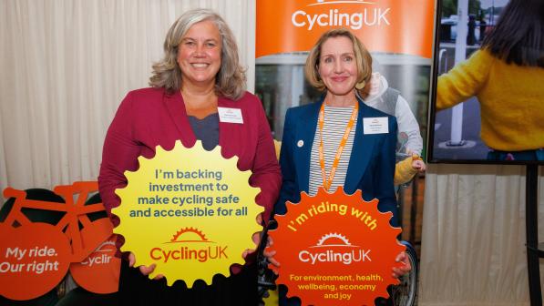 Two women are standing next to each other in front of Cycling UK branded boards. They are holding cog-shaped banners reading 'I'm backing investment to make cycling safe and accessible for all' and 'I'm riding with Cycling UK for environment, health, wellbeing, economy and joy'