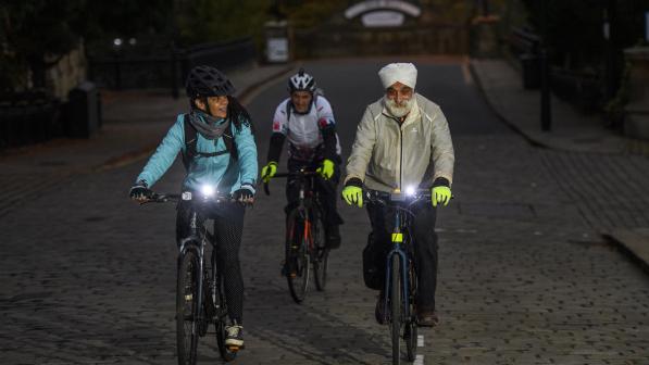 Three people are cycling along an urban street at night displaying their front white lights. Two are wearing cycle helmets, the other a turban. They're all wearing warm cycling kit.