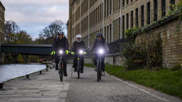 Three people are cycling along a paved canal path at either dusk or dawn. They all have bright white lights on the handlebar of their bikes