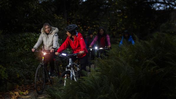 A group of people is cycling on an off-road track through trees at night. They are all wearing warm winter cycling it and have lights on their bikes