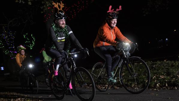 Three people are cycling on a cycle path at night. They're wearing reindeer ears and have tinsel on their bikes