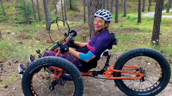 A woman is riding an orange fat trike handcycle through woodland. She is wearing purple kit and a white helmet. She has long blonde hair in a ponytail and is smiling at the camera.