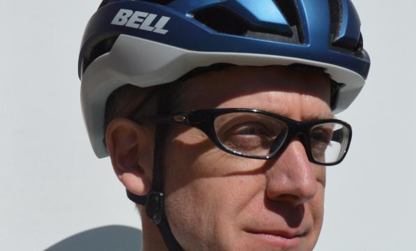 A man in glasses is wearing a blue and grey cycling helmet with the Bell logo on the side