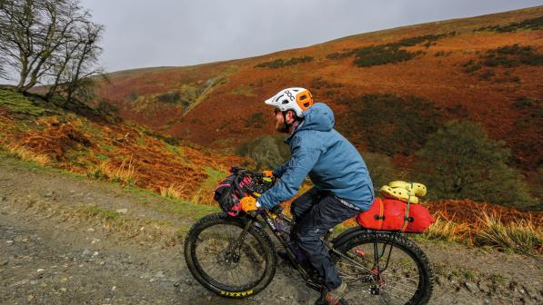 A man is riding up a slope on a gravel path through a very hilly landscape. He's wearing waterproofs and there is rain on his jacket