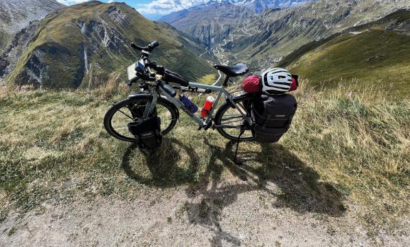 A loaded flatbar touring bike with front and rear panniers, saddlebag and top tube bag is propped up at the top of a mountain with a valley laid out behind it