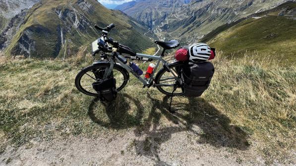 A loaded flatbar touring bike with front and rear panniers, saddlebag and top tube bag is propped up at the top of a mountain with a valley laid out behind it