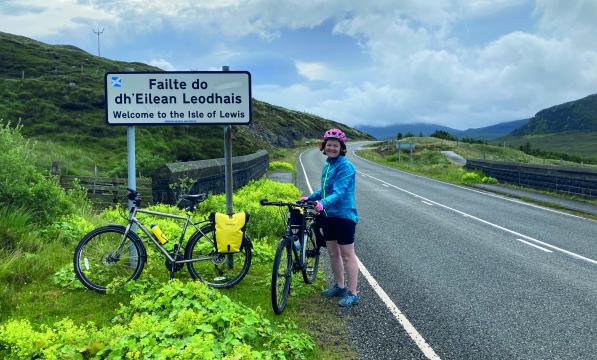 A woman is standing with her bike on the side of a road next to a road sign. She is wearing cycling shorts and helmet and a waterproof. Another bike with a packed pannier is leaning against the sign. The sign reads Failte do dh'Eilean Leodhais, with the English translation underneath: Welcome to the Isle of Lewis