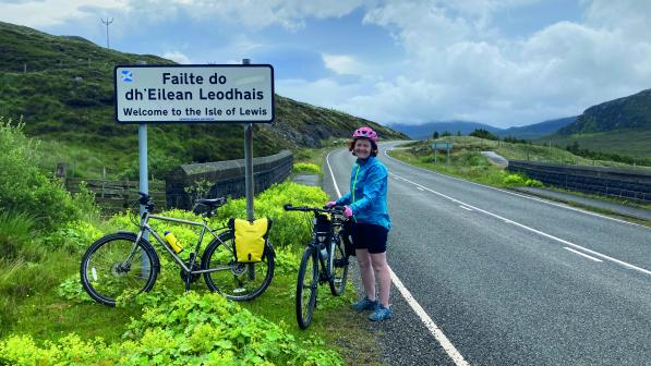A woman is standing with her bike on the side of a road next to a road sign. She is wearing cycling shorts and helmet and a waterproof. Another bike with a packed pannier is leaning against the sign. The sign reads Failte do dh'Eilean Leodhais, with the English translation underneath: Welcome to the Isle of Lewis