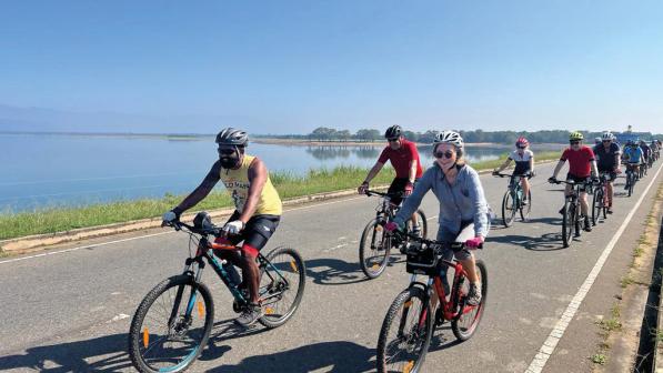 A group of people is cycling along a tarmac road in Sri Lanka. They are in a mix of cycling and non-cycling clothing, all are wearing helmets. There's a huge lake in the background.