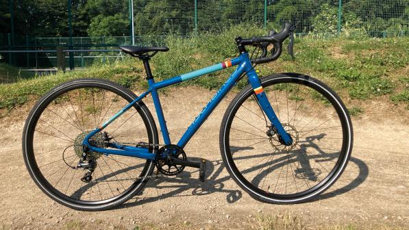 A blue gravel bike propped up on a gravel track. It has pale blue, red, yellow and orange rings painted around the frame