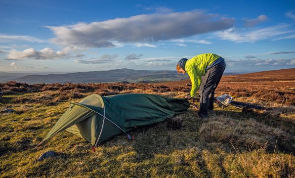 A man is setting up a bivvy bag in the countryside. He is wearing waterproof trousers and bright yellow waterproof jacket. A bike is lying on the ground in the background