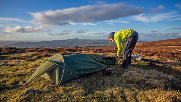 A man is setting up a bivvy bag in the countryside. He is wearing waterproof trousers and bright yellow waterproof jacket. A bike is lying on the ground in the background