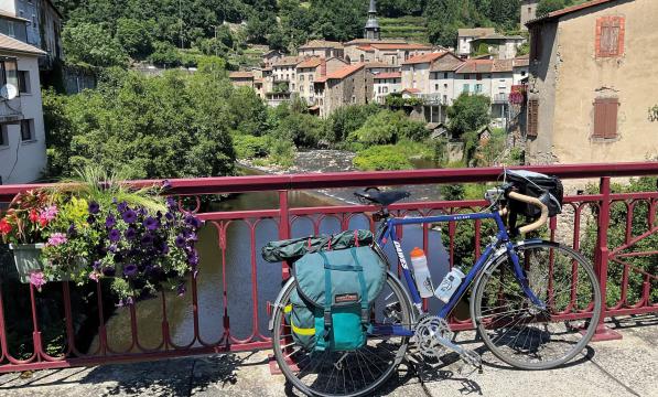 A blue touring bike is leaning against a red fence on a bridge over a stream in a town in France