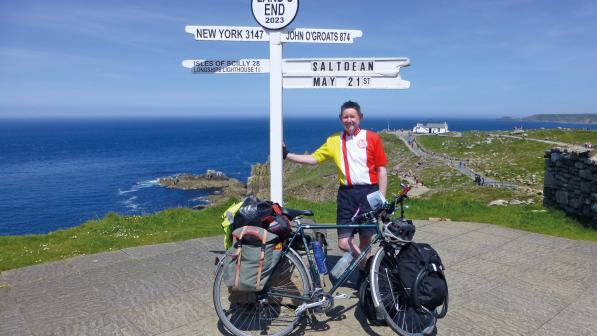  A man is standing next to the Land's End sign, which points to the Scilly Isles and John o' Groats. It also says Saltdean and the date May 21st. He is wearing shorts and a club cycling jersey. He is leaning on the sign. He has a loaded touring bike.
