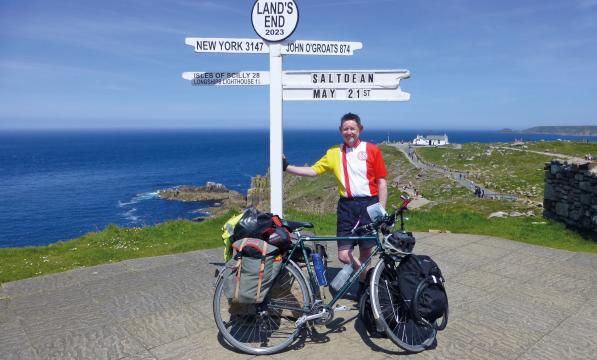  A man is standing next to the Land's End sign, which points to the Scilly Isles and John o' Groats. It also says Saltdean and the date May 21st. He is wearing shorts and a club cycling jersey. He is leaning on the sign. He has a loaded touring bike.