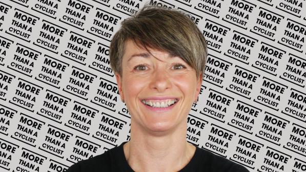A head and shoulders shot of a woman standing in front of a screen with the More Than a Cyclist logo printed all over it. She is wearing a black T-shirt with the same logo in white. She has short hair and is smiling at the camera.