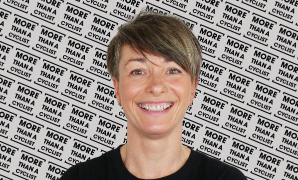 A head and shoulders shot of a woman standing in front of a screen with the More Than a Cyclist logo printed all over it. She is wearing a black T-shirt with the same logo in white. She has short hair and is smiling at the camera.