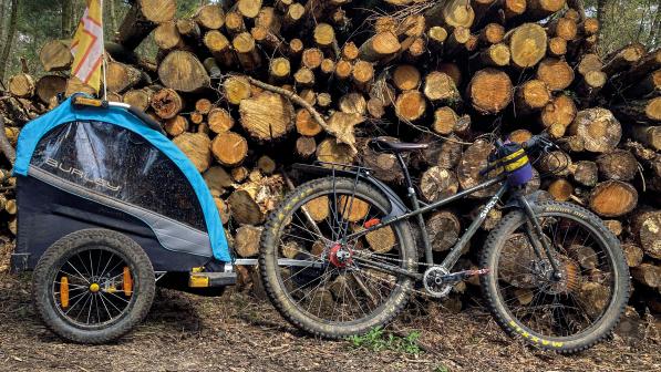 A black Surly mountain bike with a blue and black child trailer attached is leaning against a big log pile in a forest