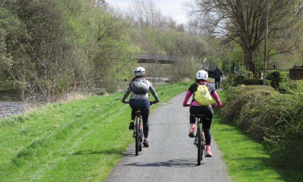 Two people are cycling along a paved path through countryside