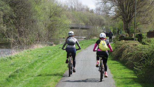 Two people are cycling along a paved path through countryside