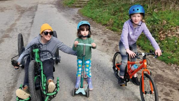 A woman on a recumbent trike is riding with two young girls, one on a scooter, the other on a bike